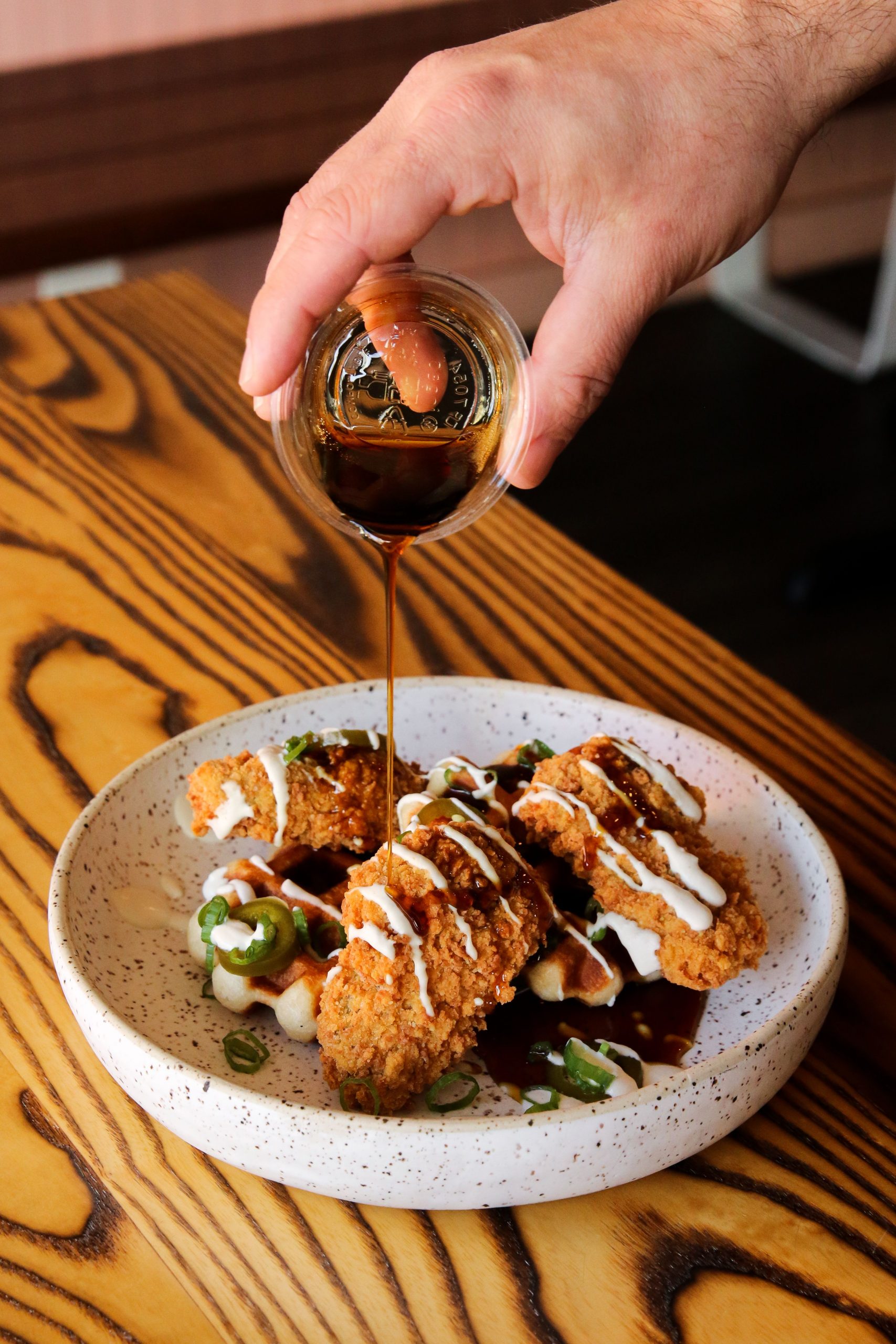 A hand pouring maple syrup on a plate of chicken and waffles 